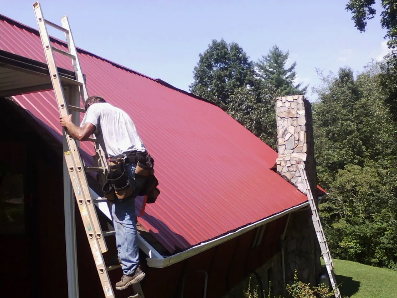 Roofer on a ladder beside a red metal roof and stone chimney — Soffit & Fascia Repair by Roofing Brothers in Macedon